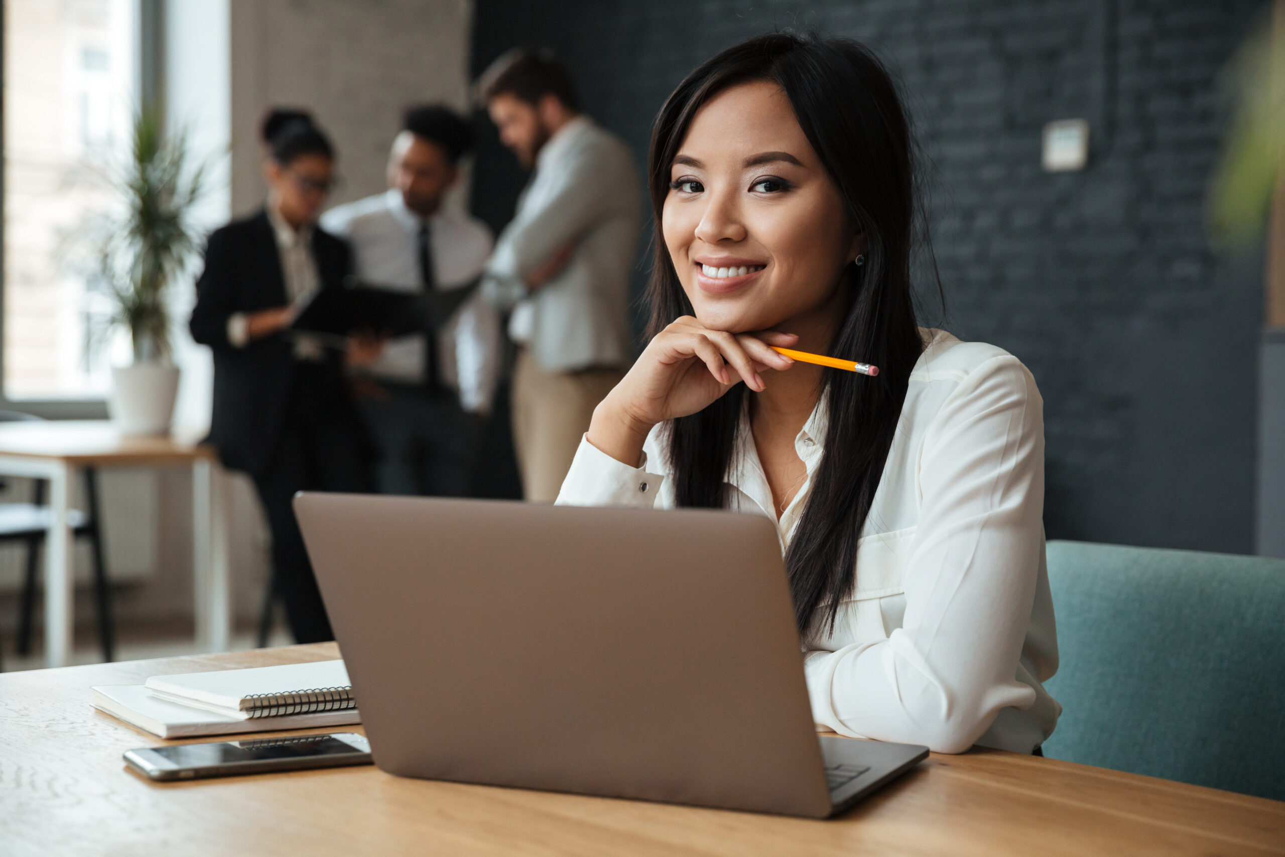 Picture of smiling young asian business woman sitting indoors using laptop computer. Looking camera.