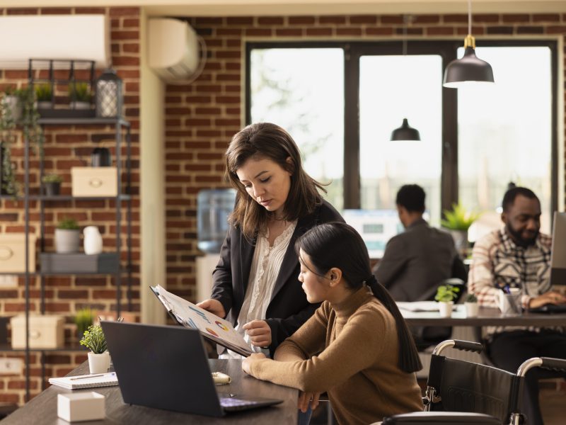 Confident female employees concentrating on business analytics, using clipboard to review financial documents. Caucasian woman standing and having a discussion with asian coworker in wheelchair.